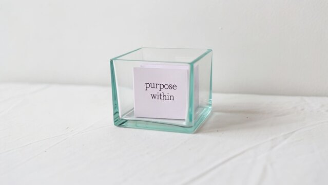 Clear glass box with a reminder card inside displaying the word "purpose" on a soft white surface with subtle natural lighting