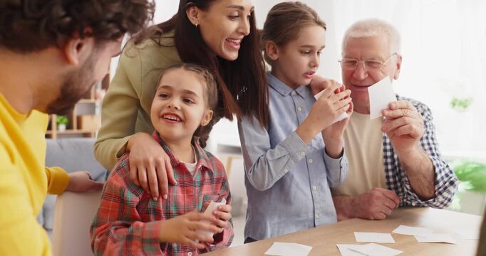 Family grandparents parents children play boardgame together. Smiling girls and kids hold cards, learning and cooperating at a home table indoors. Clear teamwork and family bonding concept.