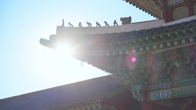 Sunlight illuminates intricate painted eaves and traditional roof tiles at Gyeongbokgung Palace in Seoul, South Korea. Guardian figurines stand along the ridge with vibrant dancheong patterns visible.