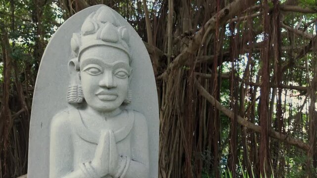 A stone carving of a figure with hands pressed together in prayer stands before a large banyan tree in Po Nagar Temple, Nha Trang, Vietnam.