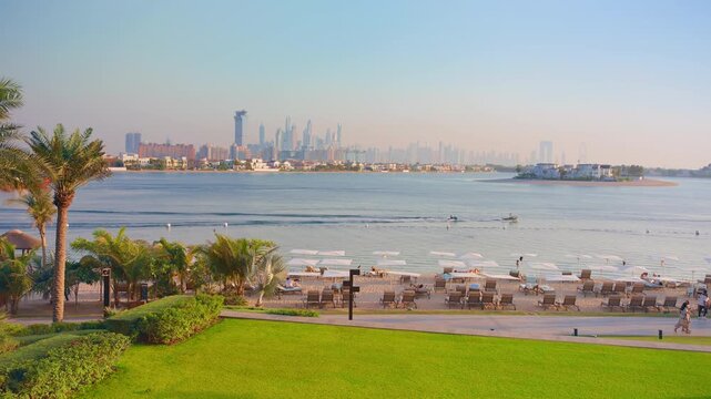 Dubai luxury resort beach with lounge chairs and umbrellas overlooking calm waters and distant city skyline. Palm trees frame the tropical garden as boats cruise by and tourists relax on sandy shore.