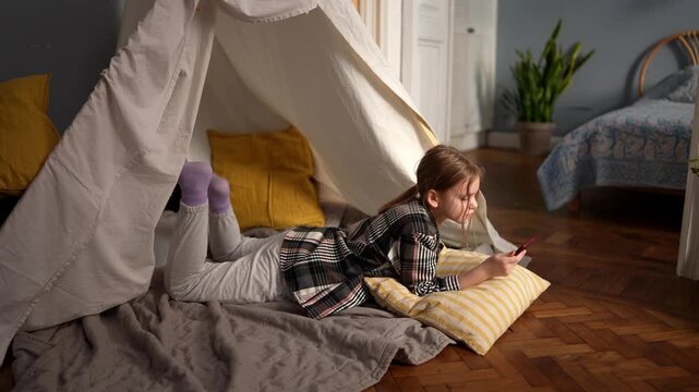 Young caucasian girl at tent entrance playing phone, kneeling then lying on stomach, purple socks and yellow pillow, wooden floor, natural window light, curious .