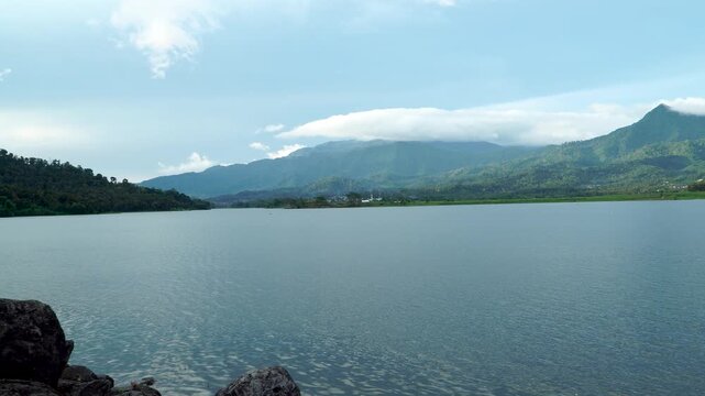 Wide landscape view of the vast Selorejo Reservoir featuring large rocks in the foreground and Mount Kelud in the background on a cloudy afternoon, Malang, East Java, Indonesia.