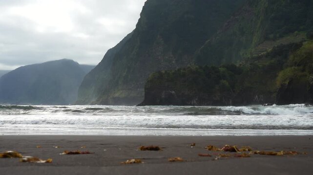 Powerful ocean waves crash onto a dark sand beach with seaweed in the foreground. Towering, lush green cliffs provide a dramatic backdrop on an overcast day. Seixal Beach, Madeira, Portugal