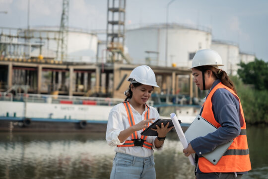 Two engineers inspect plans near industrial storage tanks. They wear hard hats and safety vests, focused on their work. Calm river flows beside the large concrete structures