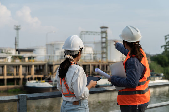 Two engineers stand by a railing, pointing at industrial facilities. They wear helmets and high-visibility vests, focused on plans. Sunlight glints off metal structures, casting soft shadows