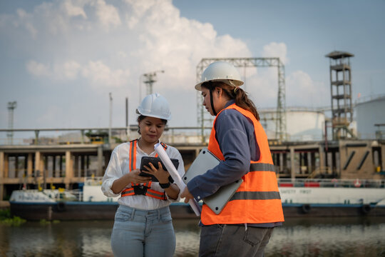 Two professionals in safety gear discuss plans by a dock. One holds a tablet, the other a clipboard. They stand near industrial structures and calm waters