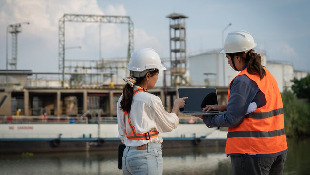 Two engineers stand near an industrial waterway. They wear hard hats and safety vests. One points at a tablet, reviewing data. The other listens, focused on the task. Background shows tall structures