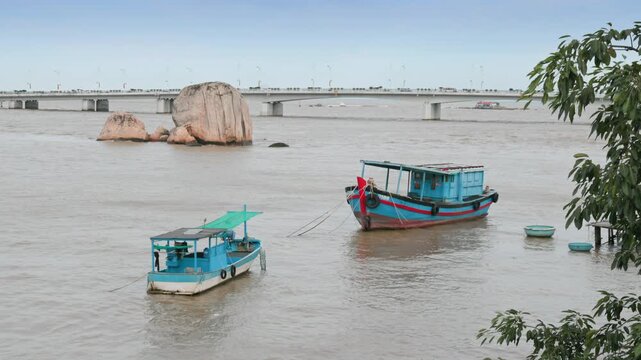 Two blue fishing boats float on the murky water near large rock formations and a bridge in Nha Trang, Vietnam.