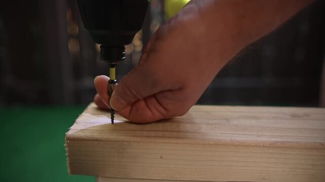 Man driving screw into wooden board using electric screwdriver tool creating process of assembly and fastening materials during construction and carpentry work