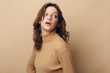 Fototapeta premium Young woman with curly hair wearing beige turtleneck looks sideways with surprised expression. Studio portrait on neutral background. Concept of curiosity, emotion, and casual fashion.