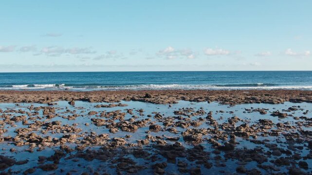 Cinematic drone shot flying forward over a rocky shore with tide pools toward the ocean horizon. Clear blue sky and calm waves in a tropical landscape.
