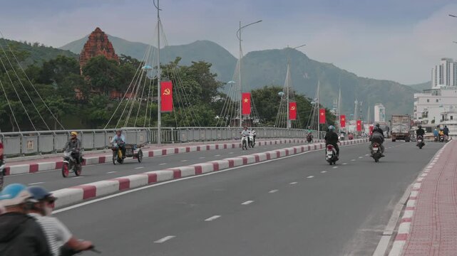 Motorcyclists ride across a bridge in Nha Trang, Vietnam, with the Cham Po Nagar towers visible in the background.
