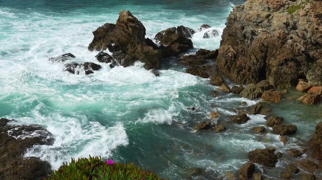 High angle slow motion view of strong ocean waves breaking on a rocky coast. Beautiful clear blue water, white foam, and brown rocks create a dramatic and powerful mood. Porto Covo, Setubal, Portugal