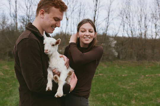 A couple outdoors holds a playful goat kid, sharing a tender moment in a grassy field, capturing lifestyle, happiness, nature, and warm bonding in a rustic countryside scene. Sunny day.