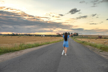 Obraz premium Young woman in blue floral dress stands on dirt road beside golden wheat field at sunset, with distant houses and cloudy sky in the background
