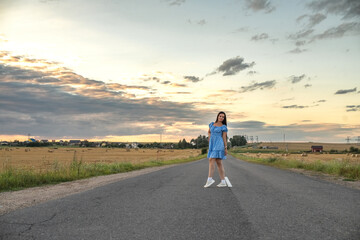 Obraz premium Young woman in blue floral dress stands on dirt road beside golden wheat field at sunset, with distant houses and cloudy sky in the background