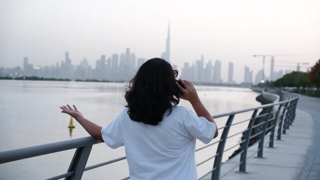 Woman walking along Dubai waterfront talking on smartphone with Burj Khalifa skyline, 4K video