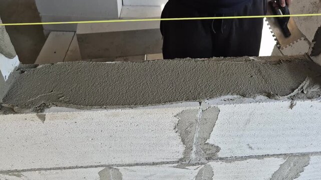 A person uses a metal trowel to spread gray mortar across a row of white AAC blocks beneath a yellow leveling line.