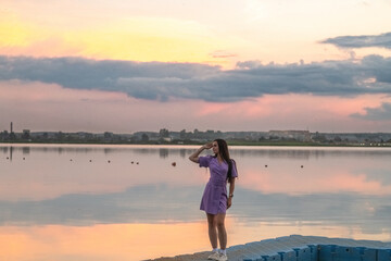 Fototapeta premium Young woman in purple dress stands on blue dock by calm lake at sunset, with dramatic clouds and colorful sky reflecting in the water