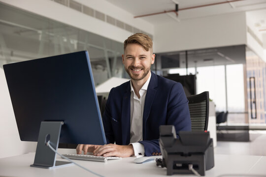 Portrait of smiling confident businessman in suit sits at desk in modern office, looking at camera while typing on computer keyboard. Corporate customer services, IT support, or administrative tasks