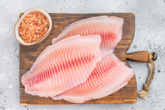 Raw tilapia fillets on wooden cutting board with sea salt, fresh freshwater fish from aquaculture on light background.