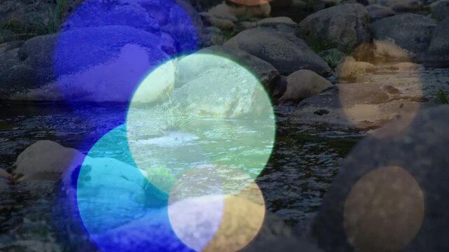 Light overlays starting to drift, colored bokeh moving across creek, revealing stones to show mood