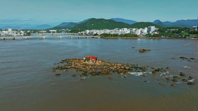 A small, colorful shrine of Hon da Chu sits on a rocky outcrop in the Cai River, Nha Trang, Vietnam, with the city skyline and mountains in the background.