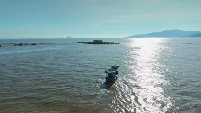 A lone fishing boat sails on the calm waters near Hon da Chu, Nha Trang, Vietnam. The sun glints off the sea as the boat heads towards the shore