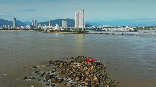 A small island of Hon da Chu with colorful buildings sits in the Cai River in Nha Trang, Vietnam, with the city skyline and mountains in the background.