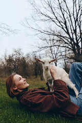 Fototapeta premium Relaxed woman lies on grass in a park, stroking a playful goat kid, enjoying outdoor life and gentle connection with nature, candid moment of simple countryside joy