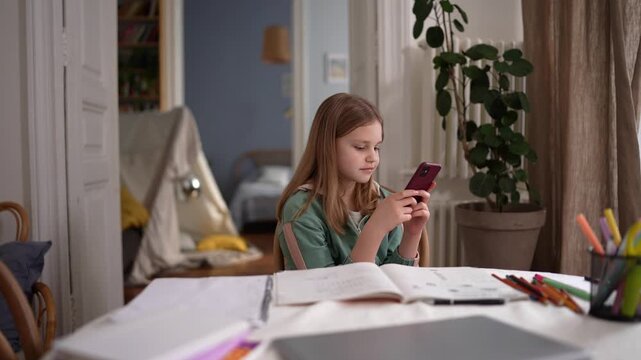 White girl checking phone during homework at table, open books and colored pencils scattered, laptop in foreground and plant by window, intent expression .