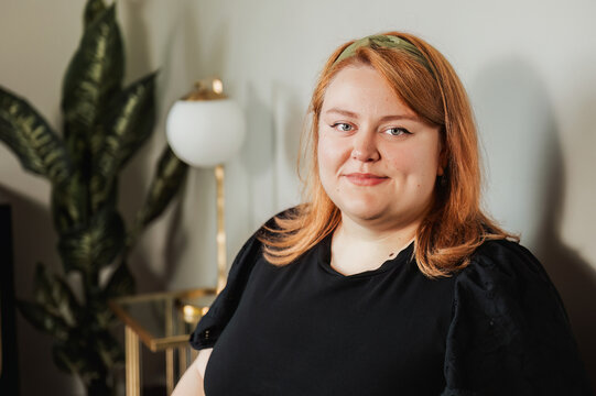 Woman with soft smile, houseplant and lamp in background indoors