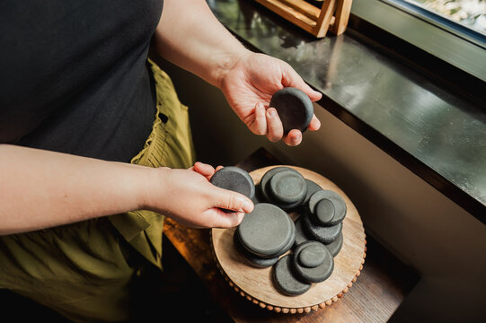 Hands holding and sorting massage stones on wooden tray by window