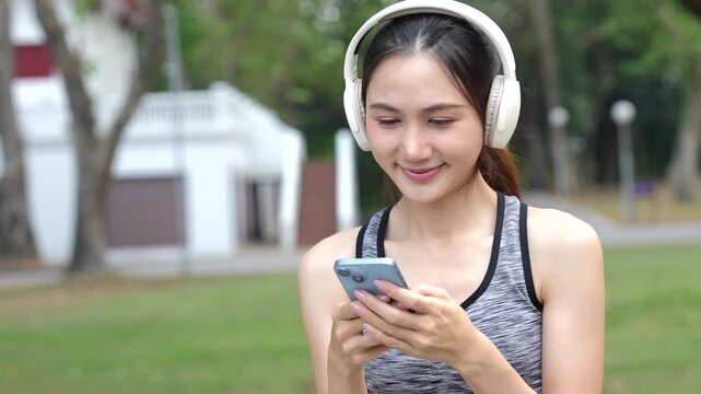 A young woman wearing headphones reads a notebook in a park, enjoying music and learning outdoors after exercise in a calm natural setting.