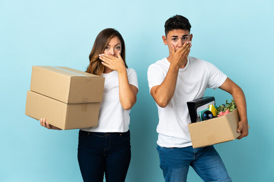 Young couple moving in new home among boxes isolated on blue background covering mouth with hands for saying something inappropriate