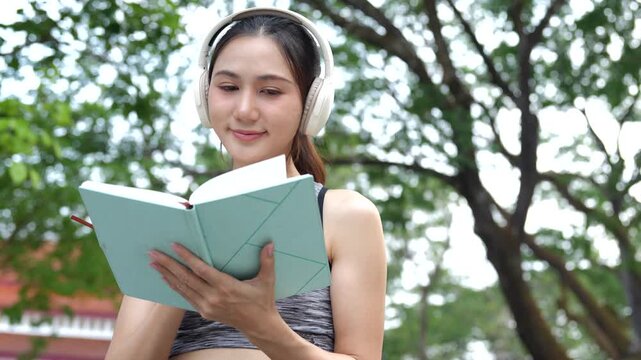 A young woman wearing headphones reads a notebook in a park, enjoying music and learning outdoors after exercise in a calm natural setting.
