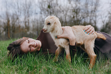 Naklejka premium Smiling woman lying on grass in a rural field, embracing a goat kid. Warm natural light highlights gentle bonding, lifestyle moment, animal companionship, and outdoor wellness.
