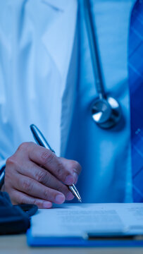 Male doctor wearing white lab coat stethoscope writes medical document upon clipboard inside blue clinical office setting performing daily healthcare duties hospital staff clinic desk patient record.