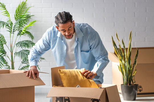 Happy young man with braids unpacking cardboard boxes with pillows in bright new apartment, moving day concept, positive lifestyle