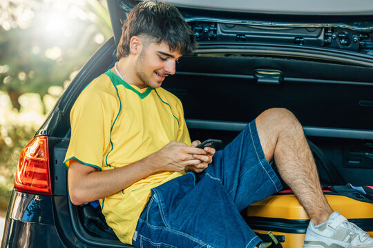 smiling young man in a summer outfit is sitting in the open trunk of a car, using his smartphone while traveling with a yellow suitcase on a sunny summer day