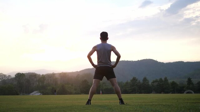 Cinematic back view of a male athlete performing a standing full body stretch in a park, scenic mountain landscape at sunset background, wellness and healthy lifestyle concept