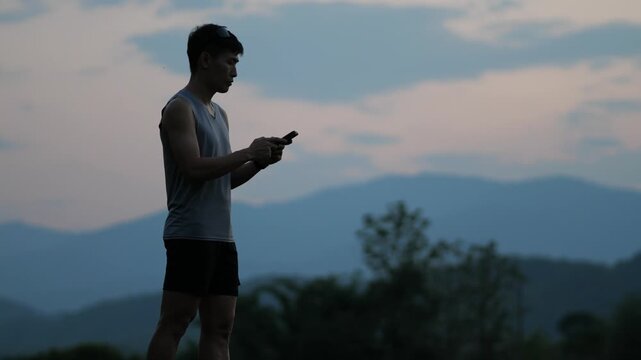 Cinematic shot of a male athlete standing and using a smartphone during a workout break, beautiful mountain view background at sunset, modern fitness and connectivity concept