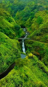 Lush green hills cradle a cascading waterfall, bisected by a winding road and quaint bridge, creating a serene and picturesque natural landscape.