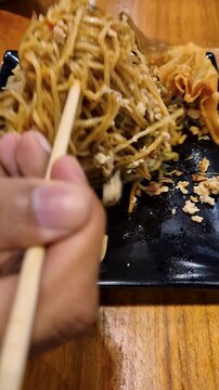 A savory noodle dish with crispy chips, spices, served on a black plate on a rustic wooden table.