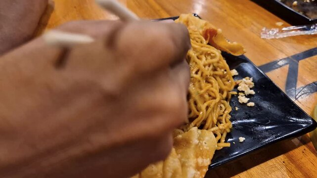 A savory noodle dish with crispy chips, spices, served on a black plate on a rustic wooden table.