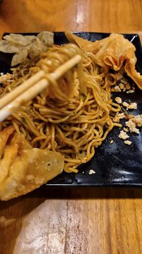 A savory noodle dish with crispy chips, spices, served on a black plate on a rustic wooden table.