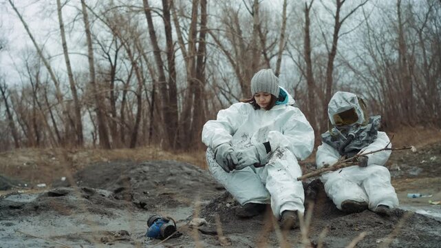 Two people in protective suits sit on muddy clearing under bare trees, sharing ration and checking radio, muddy boots and scattered gear, exhausted posture and quiet camaraderie in cold overcast
