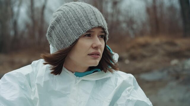 White female scientist in winter shore wearing knit beanie and protective coverall, surveying abandoned riverside with tense expression, cold wind whipping hair, muddy bank and frostcovered grass,
