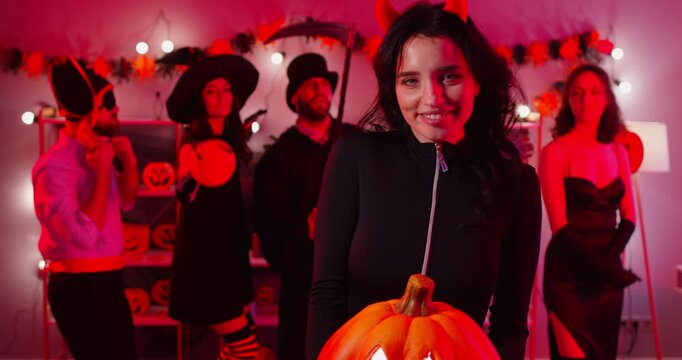 Young happy woman in black devil costume dancing with friends at Halloween party holding pumpkin in hands. People group having fun celebrating holiday at home. Celebration concept. 4k video.